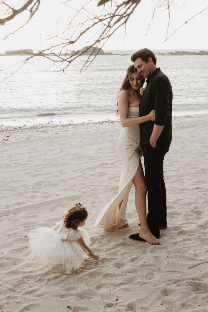 Bride and groom embrace on beach while flower girl plays in sand, elopement at St Regis Punta Mita