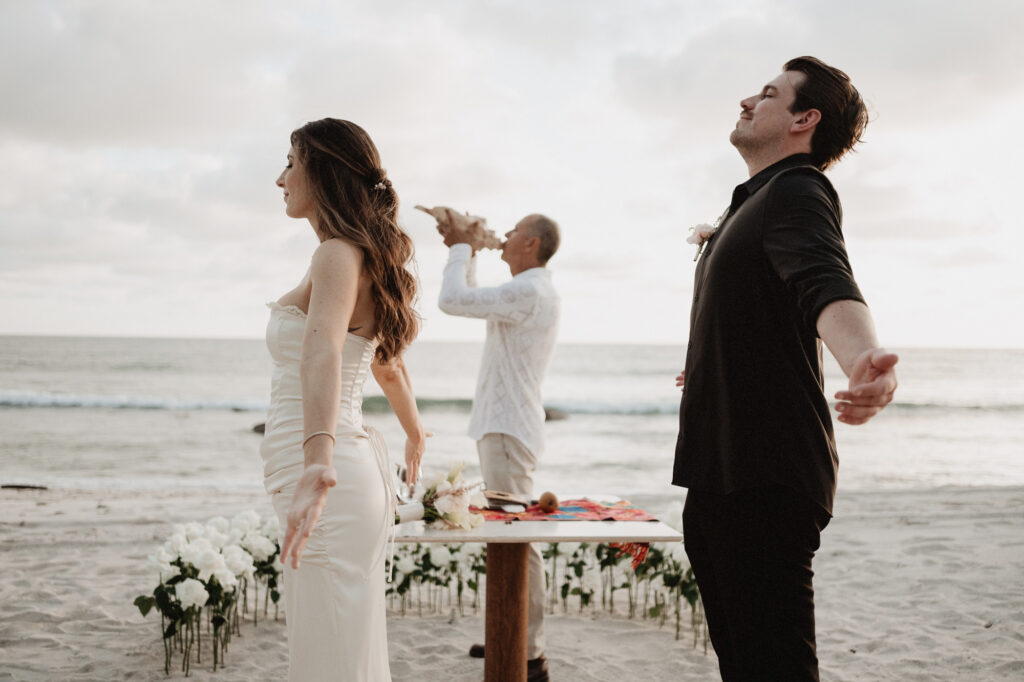 Elopement ceremony on the beach at Punta Mita, bride and groom open arms as officiant blows conch shell