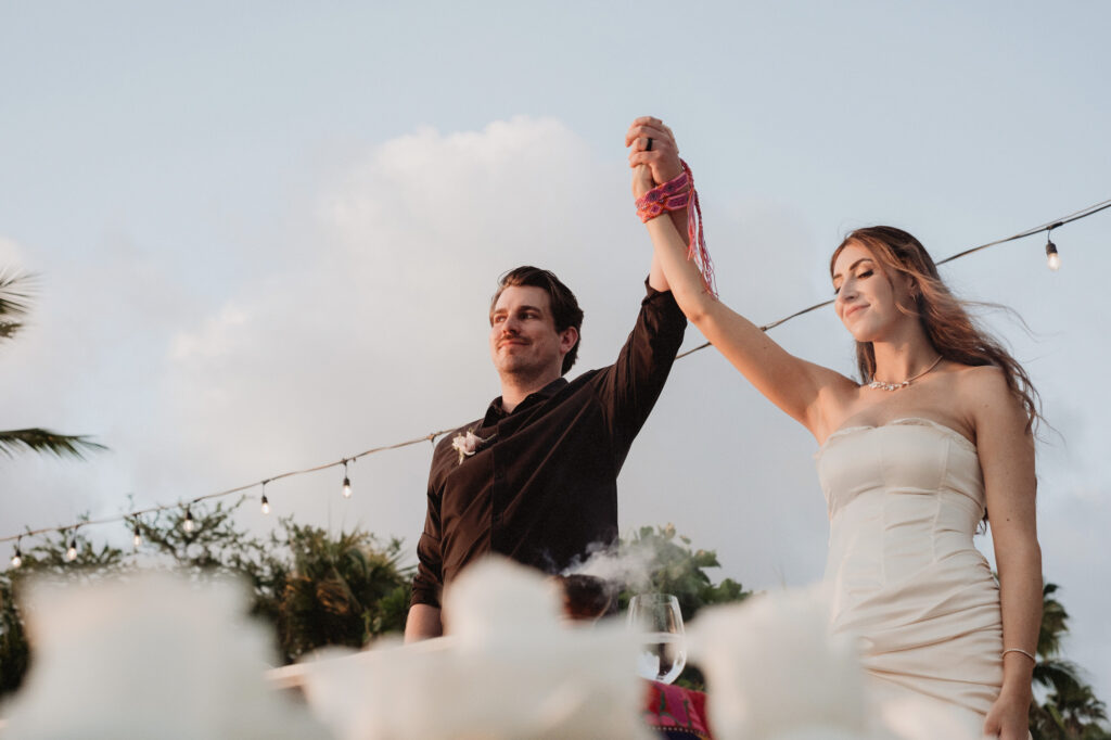 Bride and groom raise hands together at Punta Mita elopement, string lights and palm trees behind them