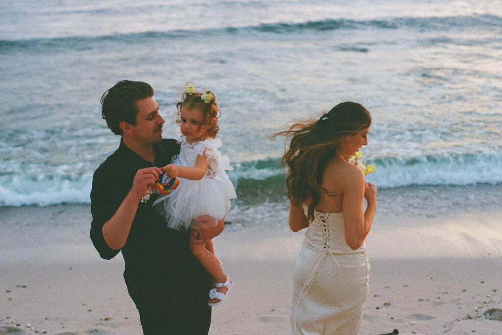 Groom holds toddler in white dress at beach, bride in lace-back gown, Punta Mita elopement