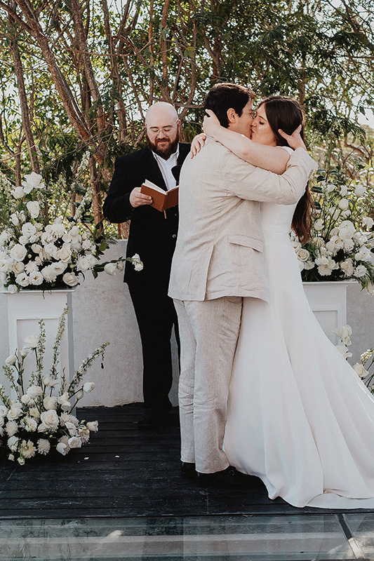 Bride and groom share first kiss at outdoor wedding ceremony, Ambre & Epieces Tulum, surrounded by white florals