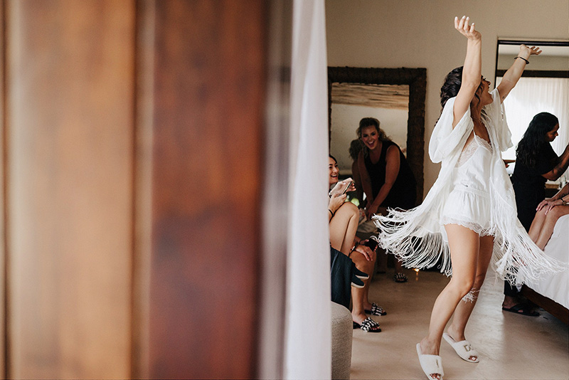 Bride dancing joyfully in white fringe robe with bridesmaids during wedding prep at Papaya Playa Project Tulum