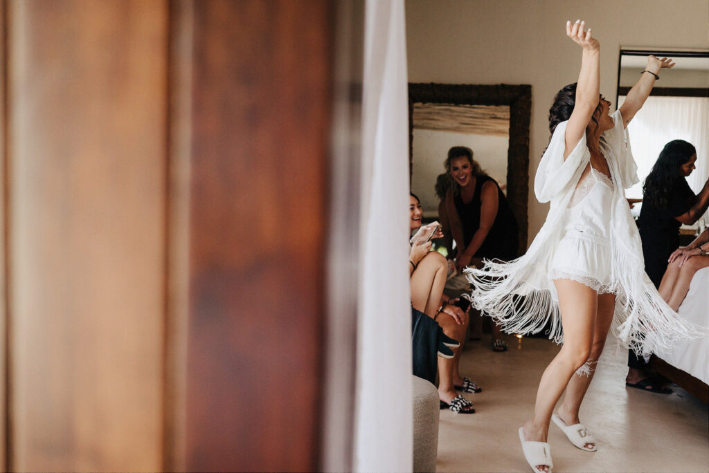 Bride dancing in white fringe robe with bridesmaids laughing, wedding morning prep at Papaya Playa Project Tulum