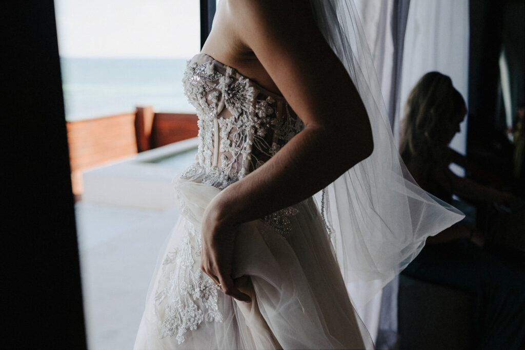 Bride in beaded lace wedding gown holding her skirt by ocean window at Papaya Playa Project Tulum
