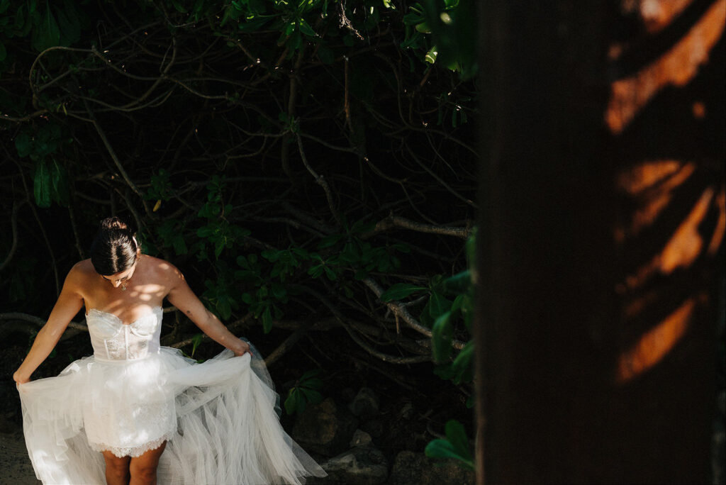 Bride in white corset gown adjusting tulle skirt among tropical trees at Papaya Playa Project Tulum wedding