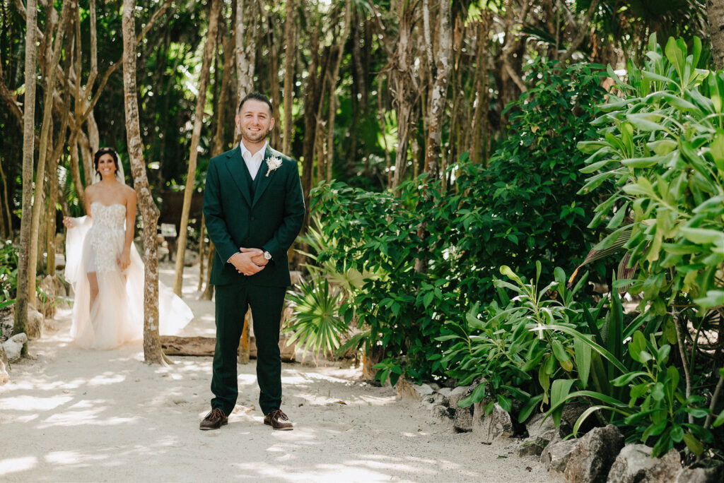 Groom in dark green suit smiles as bride approaches in tropical forest path, Papaya Playa Project Tulum wedding