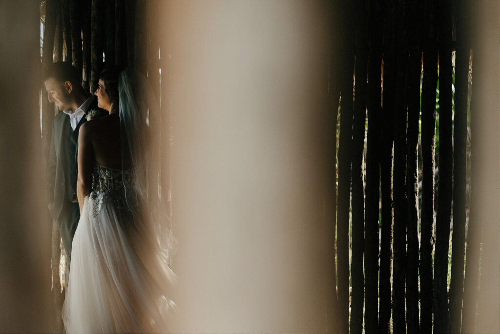 Bride and groom portrait at Papaya Playa Project Tulum, shot through rustic wooden structure, wedding day